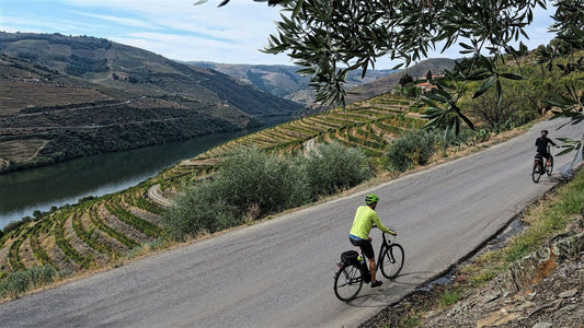 Two cyclists ride along a winding road in the Douro valley, beside terraced vineyards with a river and green hills in the background.