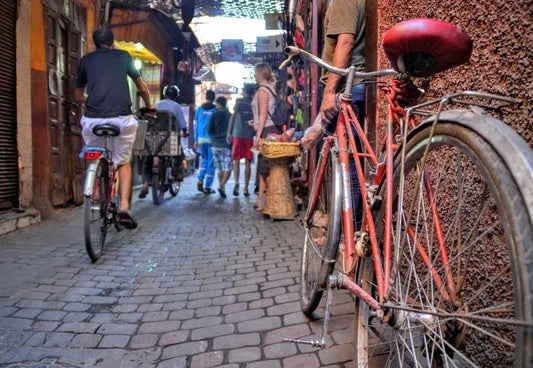 A red bicycle leans against a textured wall on a narrow cobblestone street as people walk and ride bikes through a busy, colorful market alley in Morocco.