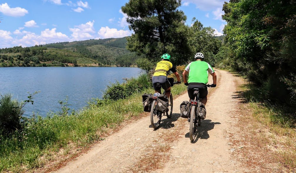 Two cyclists ride on a gravel path beside a scenic lake in Portugal, surrounded by lush green trees and hills under a partly cloudy sky.