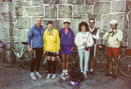 Six cyclists wearing colorful jackets, shorts, and bike helmets stand and smile in front of a stone wall with their bicycles lined up behind them.