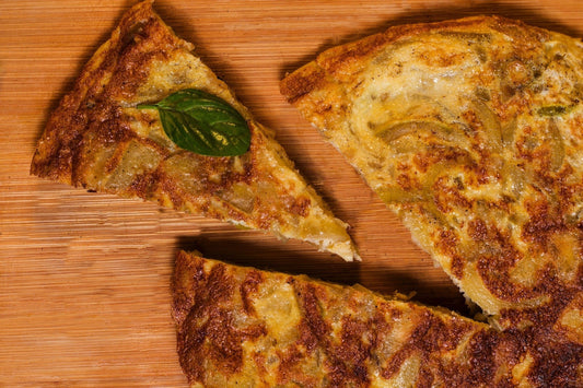 A close-up of a sliced Spanish omelette (tortilla española) on a wooden surface, with one triangular piece topped with a fresh basil leaf.