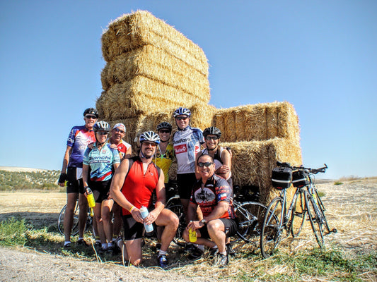 A group of cyclists in colorful jerseys and helmets pose with their bikes in front of large stacked hay bales on a sunny day in an open field.