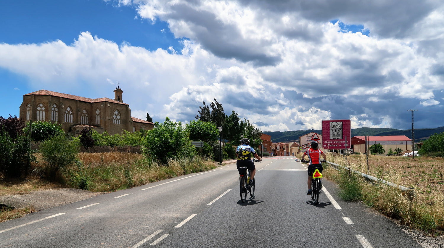A person wearing a helmet and bright shirt rides a bicycle on a paved road through a green, hilly landscape under a clear blue sky.