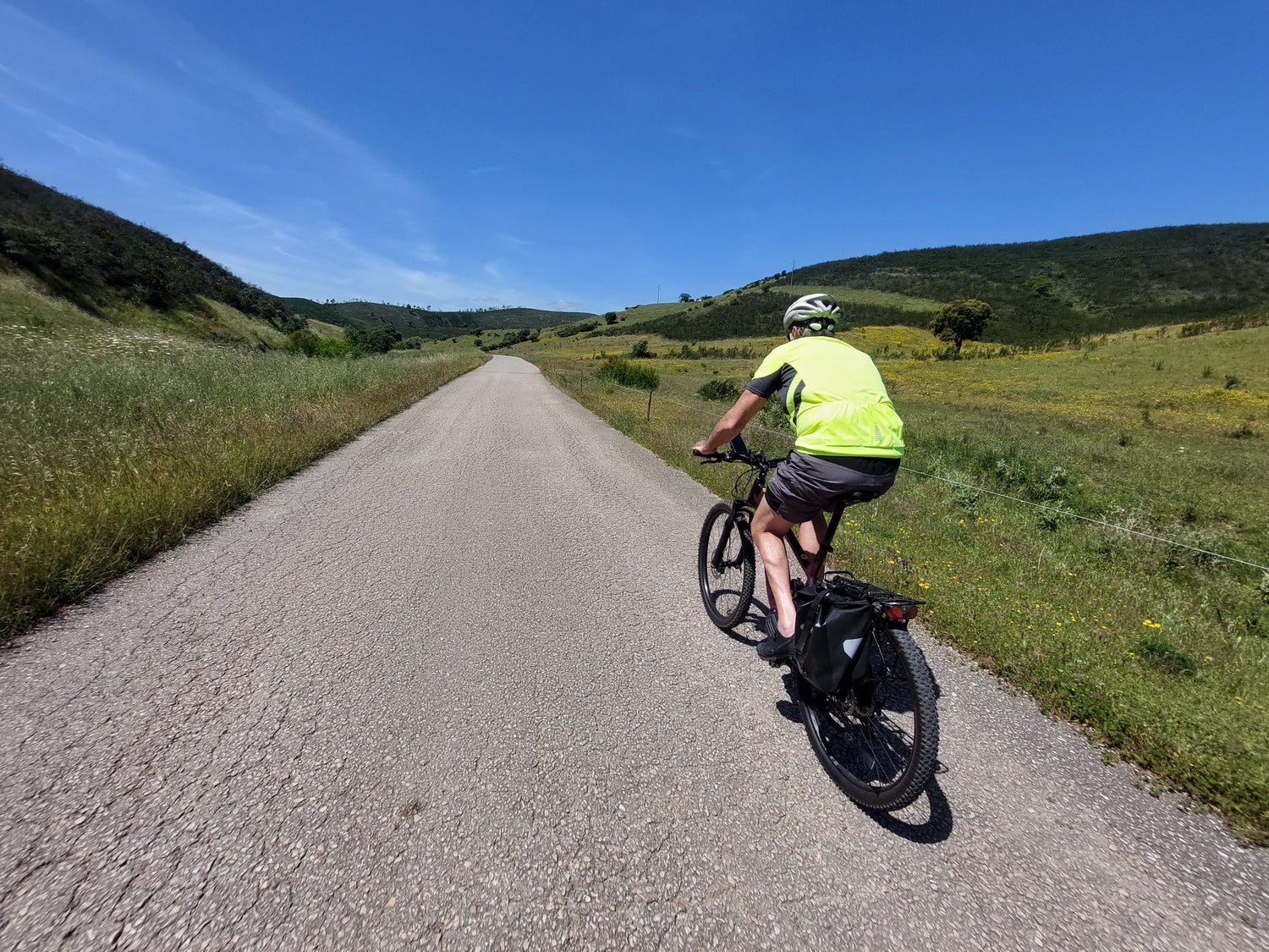 A person wearing a helmet and bright shirt rides a bicycle on a paved road through a green, hilly landscape under a clear blue sky.