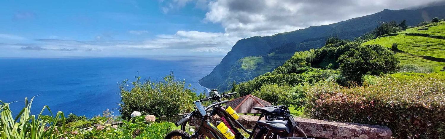 Bicycles rest against a stone wall overlooking a lush green coastal hillside in the Azores, with a vast blue ocean under a partly cloudy sky.