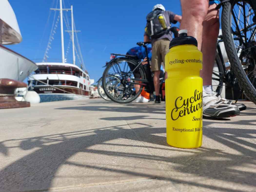 Close up of a yellow CTTC bike bottle with a cruise boat, bikes and cyclists in the background