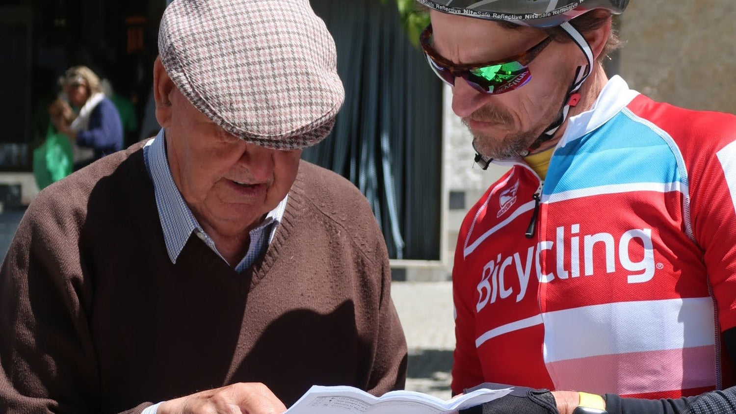 An elderly man wearing a flat cap and brown sweater points at a map while talking to a cyclist in a colorful bike jersey and sunglasses.