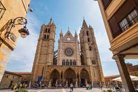 A view of the large Gothic cathedral of Leon with twin towers, a rose window, and arched entrance, set against a blue sky, surrounded by old buildings and a few people in the plaza.