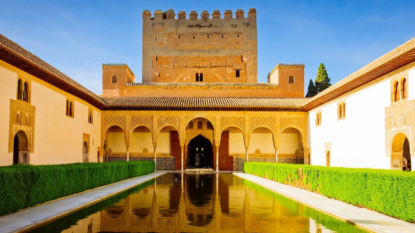 Courtyard with a central reflecting pool, decorative arches, and a large tower in the Alhambra, Spain.