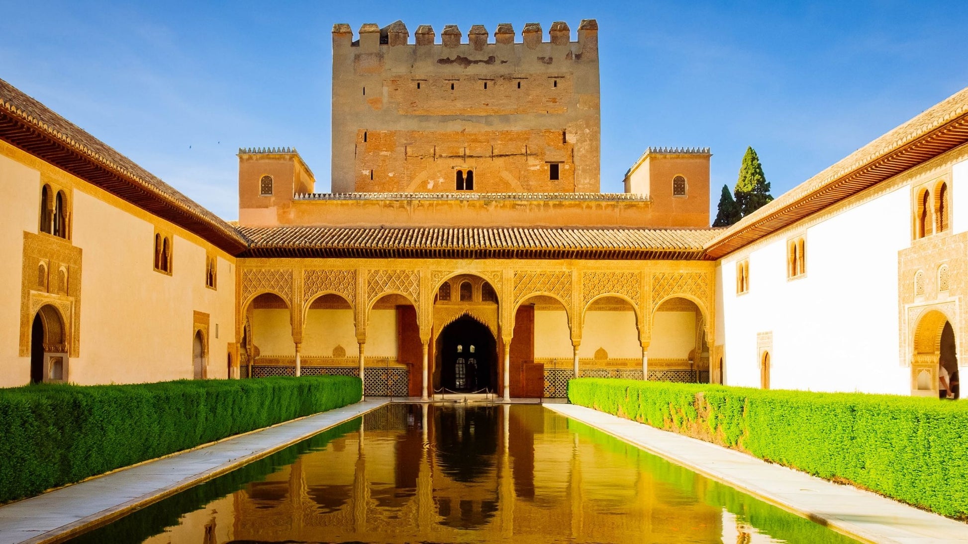 Courtyard with a central reflecting pool, decorative arches, and a large tower in the Alhambra, Spain.