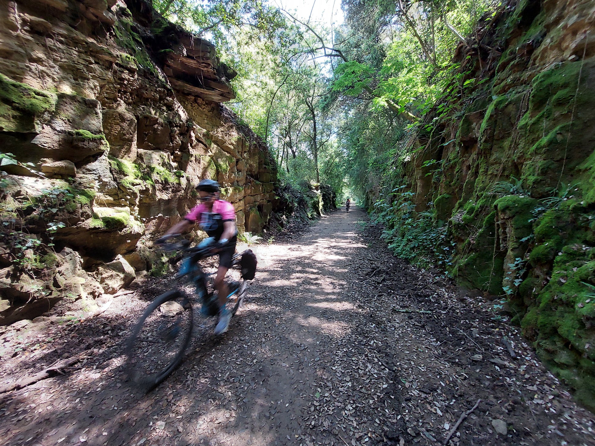A cyclist in a pink shirt rides a dirt trail past mossy rocks and lush foliage near Girona.