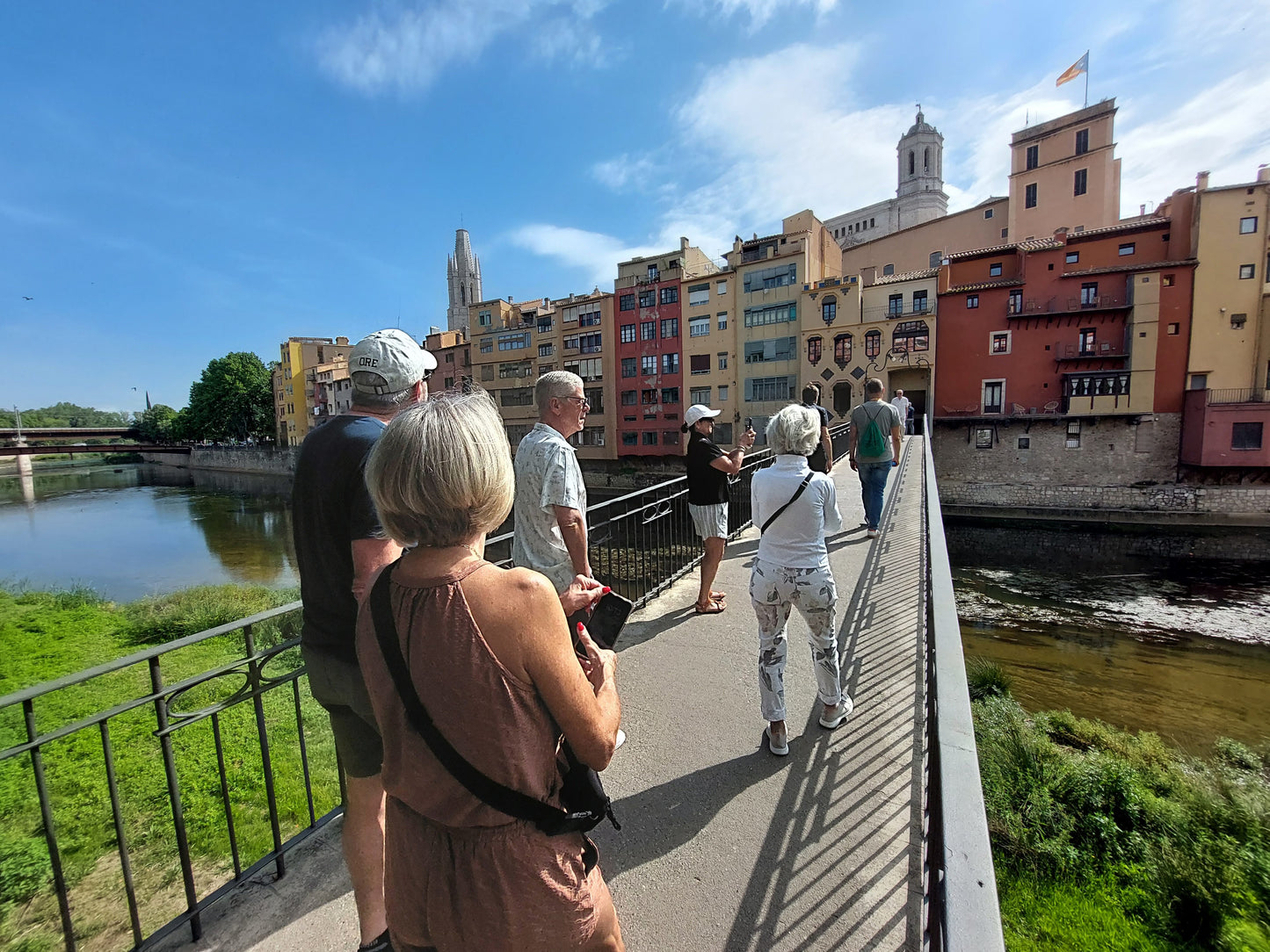 Tourists stroll across a bridge in sunny Girona, passing vibrant buildings and the historic cathedral.