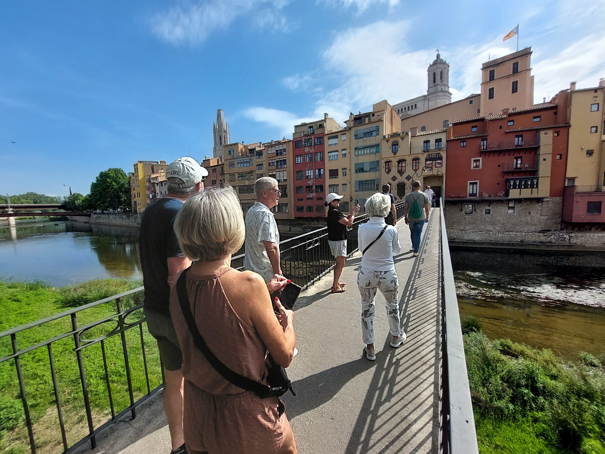 Tourists stroll across a bridge in sunny Girona, passing vibrant buildings and the historic cathedral.