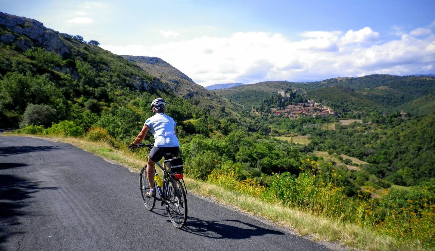 A cyclist wearing a helmet rides a bike on a paved road through the hills of Languedoc with a village in the distance.