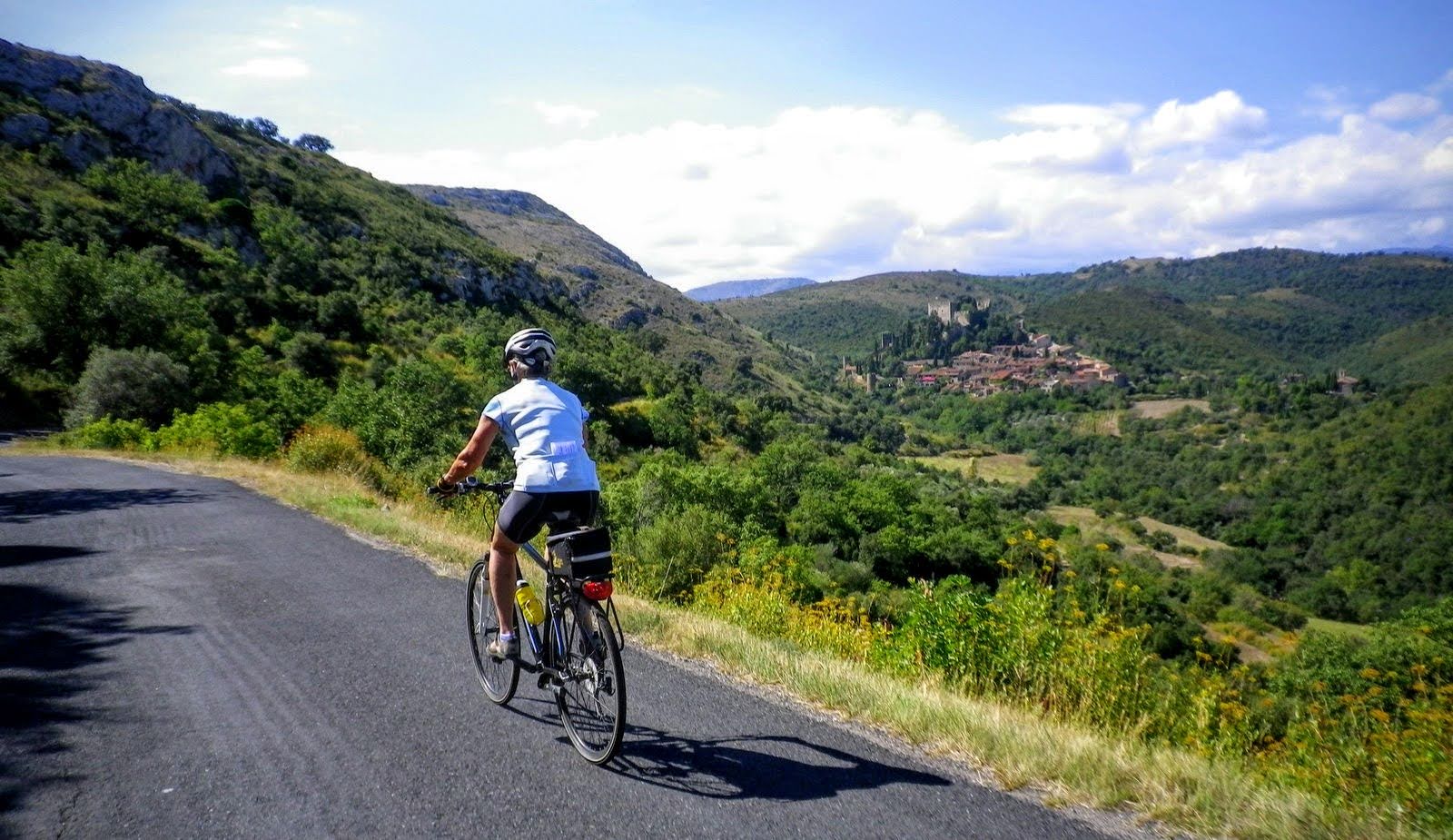 A cyclist wearing a helmet rides a bike on a paved road through the hills of Languedoc with a village in the distance.
