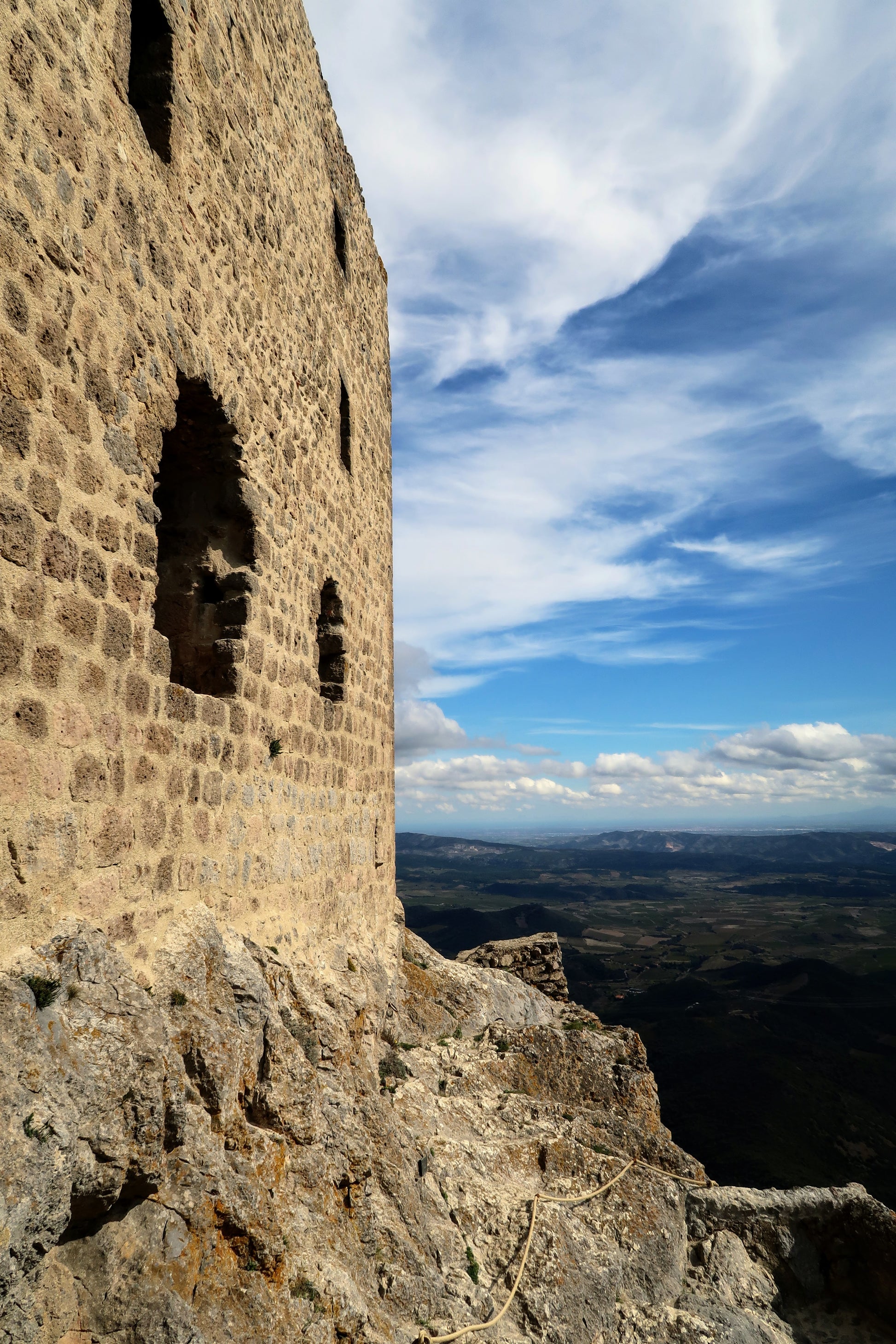 Stone wall of an old building on a rocky hilltop, overlooking a distant valley beneath a partly cloudy sky.