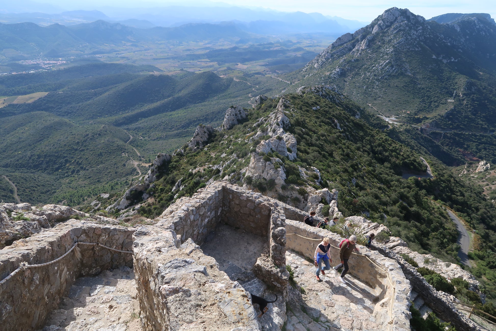 Two people climb stone steps in ancient castle ruins atop a mountain in Languedoc, lush valleys and distant hills around them