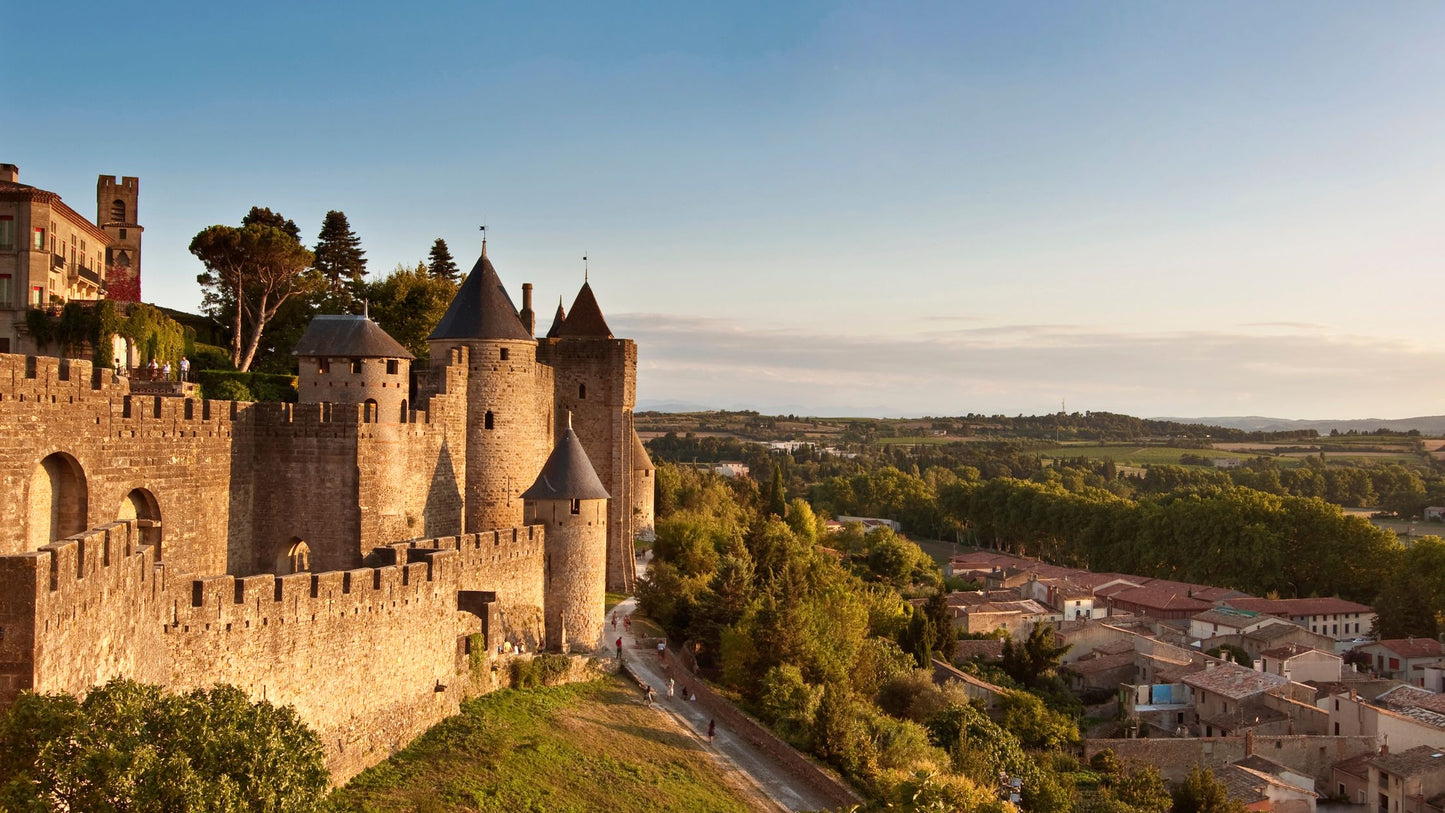 Medieval stone fortress with towers and crenelated walls overlooks a town and green countryside under a clear sky.