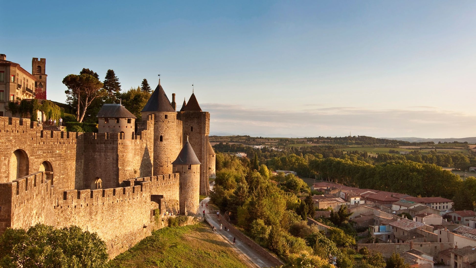 Medieval stone fortress with towers and crenelated walls overlooks a town and green countryside under a clear sky.