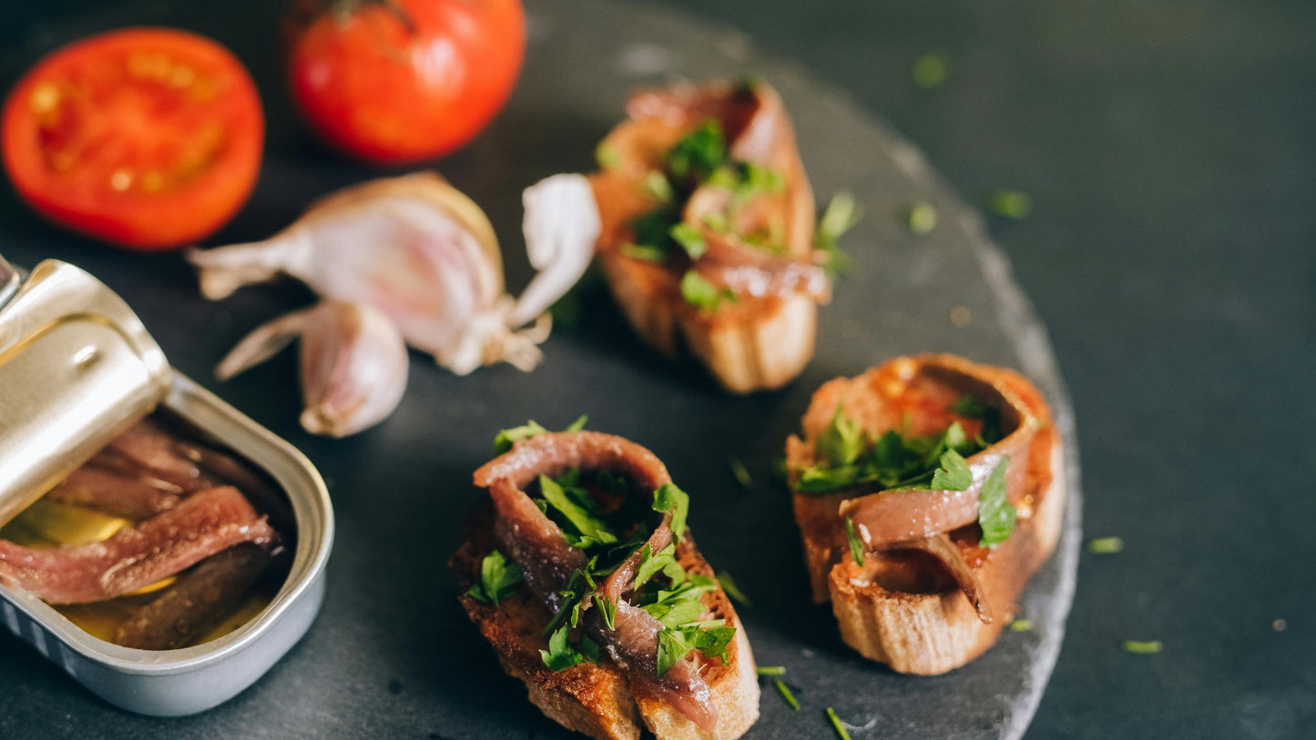 Three pieces of bruschetta with anchovies and fresh herbs, paired with a tin of anchovies, garlic, and tomatoes.