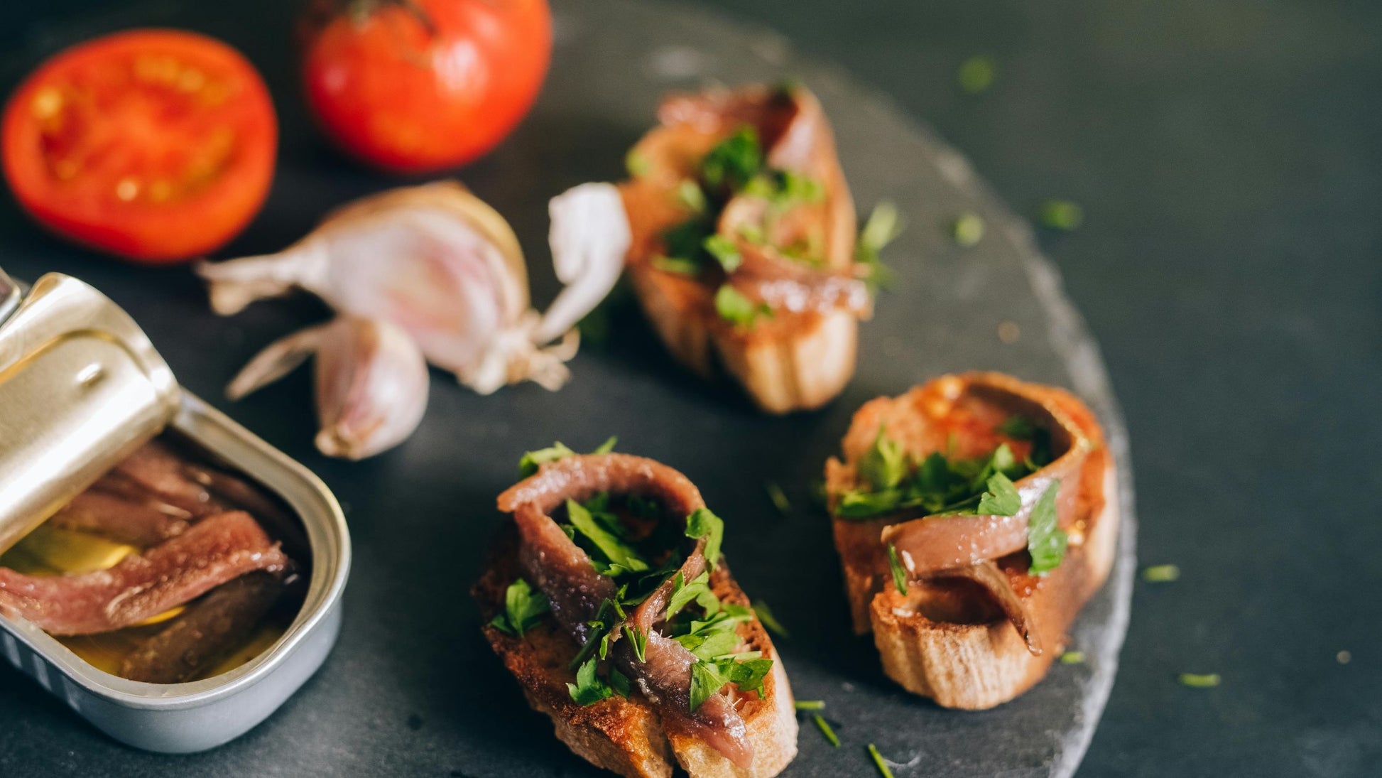 Three pieces of bruschetta with anchovies and fresh herbs, paired with a tin of anchovies, garlic, and tomatoes.