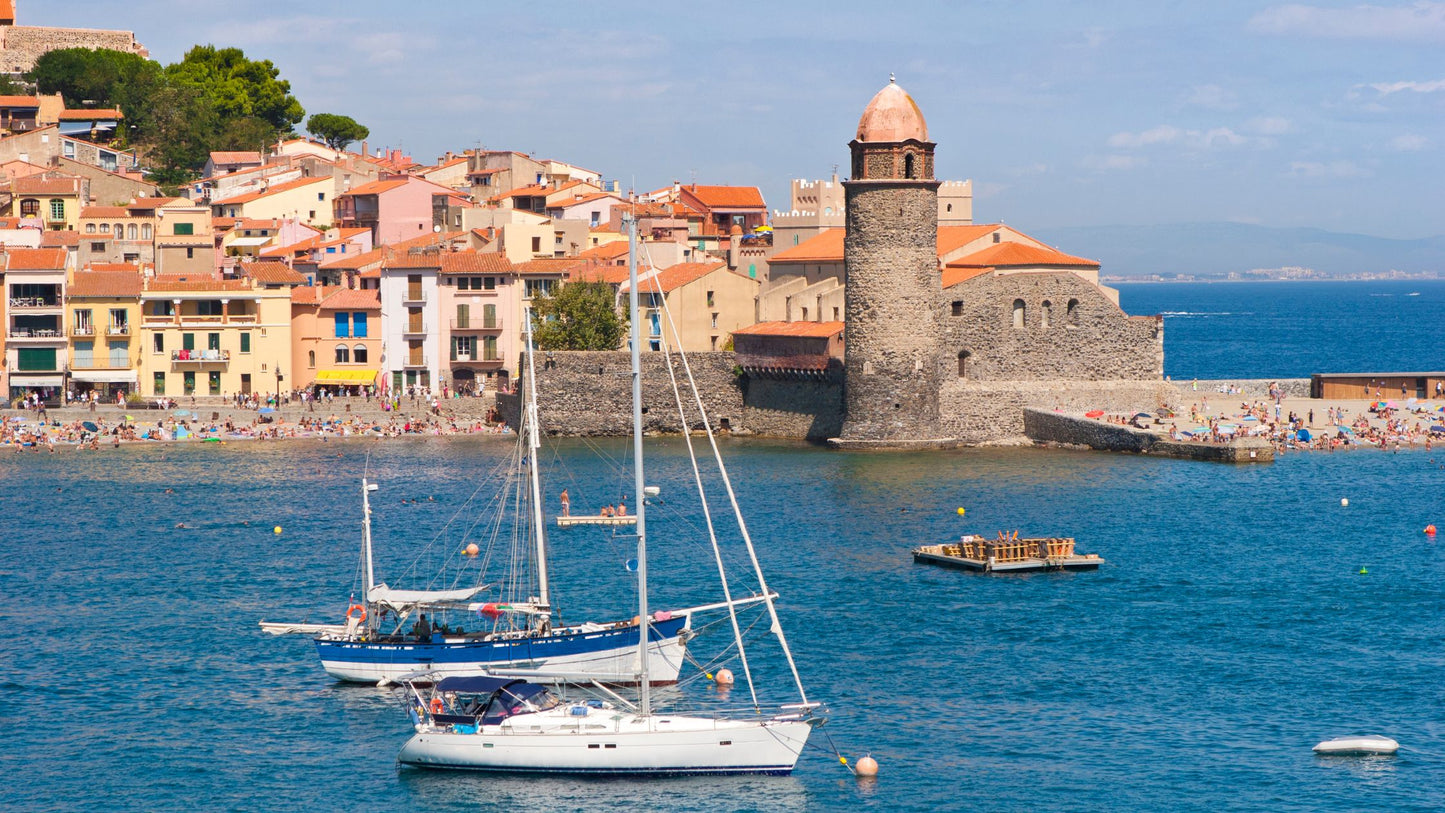 Sailboats anchor in a blue harbor beside a historic stone church and vibrant waterfront, creating an idyllic scene.