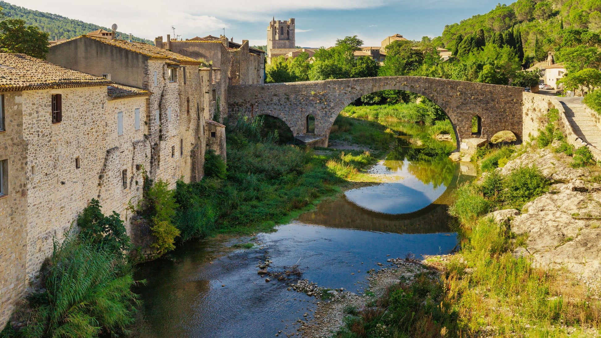 A stone bridge spans a serene river by historic buildings and lush greenery, with a church tower beyond.
