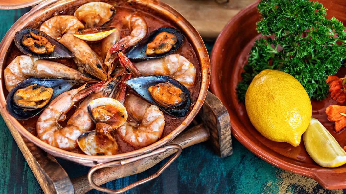 A bowl of seafood stew with shrimp, mussels, and clams, next to a plate with parsley and a whole lemon.